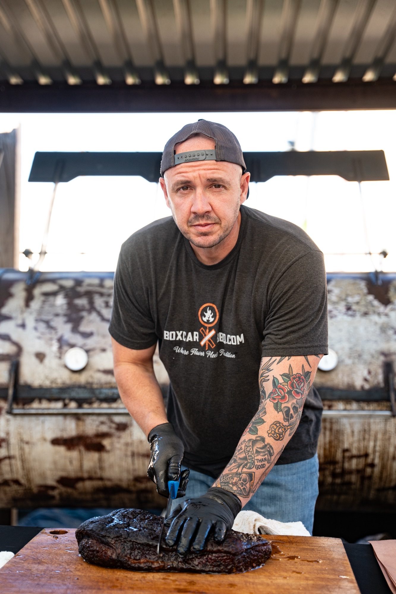 Pitmaster in a backward cap and black gloves slicing a whole brisket in front of the smoker